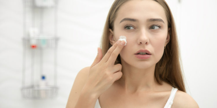 Young beautiful woman applying cream to her face and looking worried at the mirror at home bathroom. Beauty, skin care concept, lifestyle