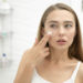 Young beautiful woman applying cream to her face and looking worried at the mirror at home bathroom. Beauty, skin care concept, lifestyle