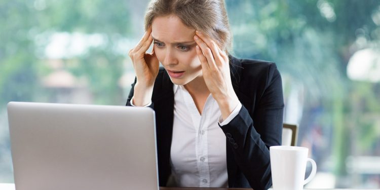 Worried young attractive businesswoman sitting at table and looking at laptop screen while touching temples. Front view with big window and blurry green view outside in background.