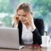 Worried young attractive businesswoman sitting at table and looking at laptop screen while touching temples. Front view with big window and blurry green view outside in background.
