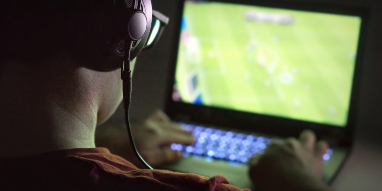 Playing video games with laptop. Young man plays online soccer or football on computer. Back view of gamer with headphones in dark or late at night. Competitive gaming, electronic sports and esports.