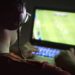 Playing video games with laptop. Young man plays online soccer or football on computer. Back view of gamer with headphones in dark or late at night. Competitive gaming, electronic sports and esports.