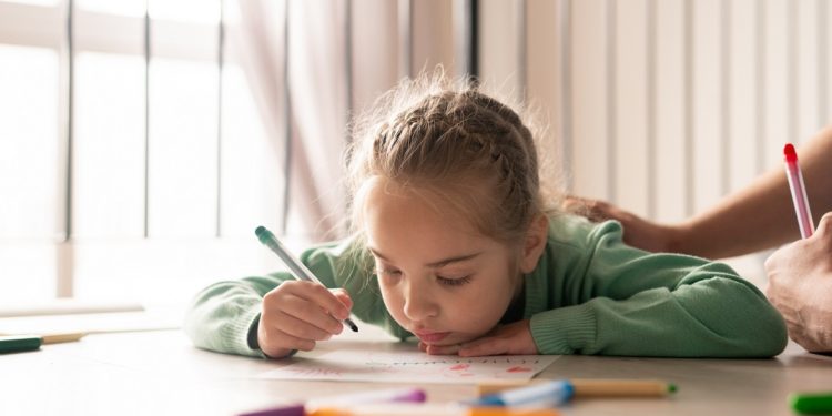 Serious lazy pretty girl with braided hair lying on floor and coloring picture with felt-tip pens while her father supporting her creativity and stroking her back
