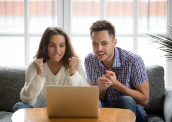 Nervous excited couple in tension clenching fists looking at laptop screen, man and woman football fans cheering supporting sport team watching championship match online together hope for win at home
