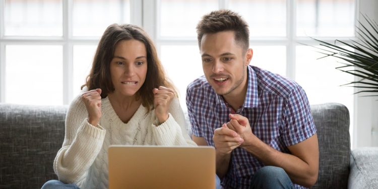 Nervous excited couple in tension clenching fists looking at laptop screen, man and woman football fans cheering supporting sport team watching championship match online together hope for win at home