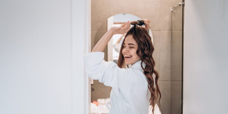 Beautiful smiling woman curling long hair using curling iron in the bath