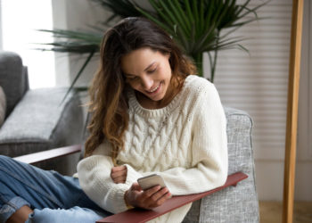 Smiling happy millennial woman holding smartphone relaxing on armchair, teenage girl checking social network news online, reading texting message on cell or using app on new mobile phone at home