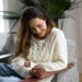 Smiling happy millennial woman holding smartphone relaxing on armchair, teenage girl checking social network news online, reading texting message on cell or using app on new mobile phone at home