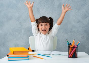 Back to school. Cute child schoolgirl sitting at a Desk in the room. The kid is learning and doing his homework. The girl is emotionally happy with her hands up.