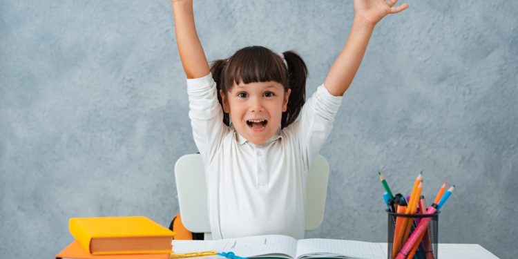 Back to school. Cute child schoolgirl sitting at a Desk in the room. The kid is learning and doing his homework. The girl is emotionally happy with her hands up.
