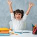 Back to school. Cute child schoolgirl sitting at a Desk in the room. The kid is learning and doing his homework. The girl is emotionally happy with her hands up.