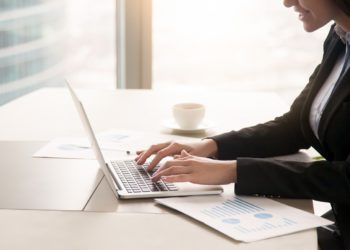 Close-up side view of serious businesswoman working at office desk on laptop, typing and looking at screen, surrounded by diagrams and calculating charts. Business, finances, economics and technology