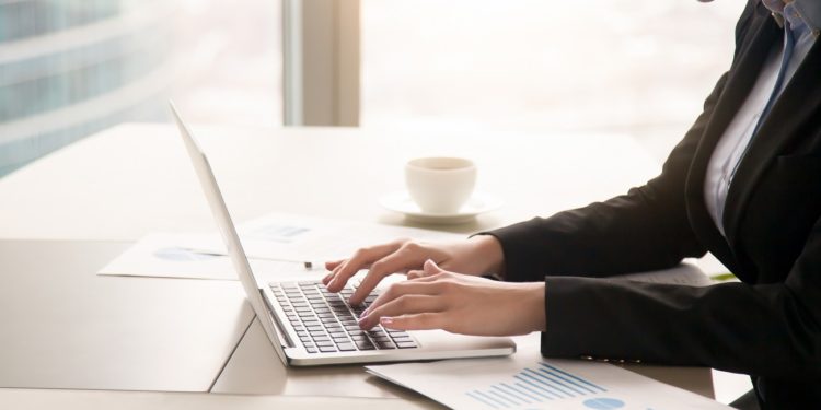 Close-up side view of serious businesswoman working at office desk on laptop, typing and looking at screen, surrounded by diagrams and calculating charts. Business, finances, economics and technology
