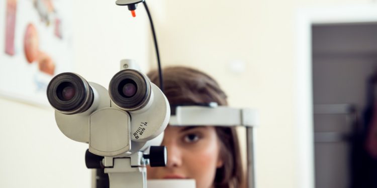 Portrait of cute caucasian female patient sitting in optometrist office, waiting for start of procedure to check her vision with microbioscope, sitting over yellow background. Ophthalmology concept.