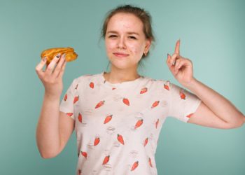 Portrait of pimply teen girl wants to eat eclair cake on blue background