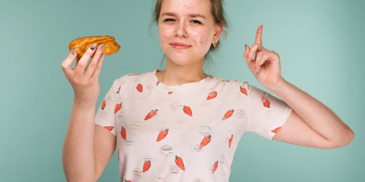 Portrait of pimply teen girl wants to eat eclair cake on blue background