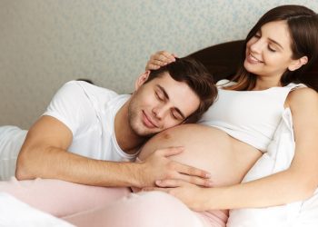 Smiling young pregnant wife lying in bed with her husband in the bedroom, man is lying on her belly