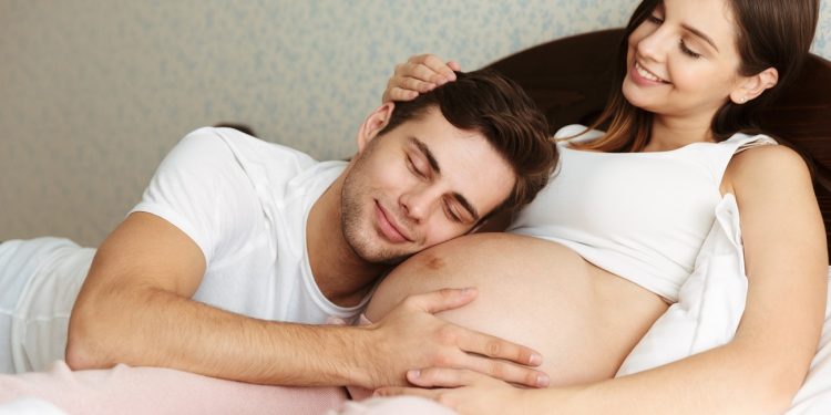 Smiling young pregnant wife lying in bed with her husband in the bedroom, man is lying on her belly