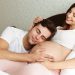 Smiling young pregnant wife lying in bed with her husband in the bedroom, man is lying on her belly
