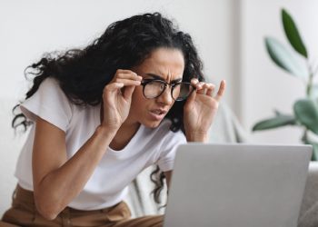 Young woman freelancer with bad eyesight using laptop, using glasses, home interior, copy space. Curly lady holding her glasses and squinting, looking at laptop screen, having vision troubles