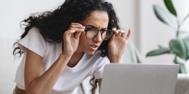 Young woman freelancer with bad eyesight using laptop, using glasses, home interior, copy space. Curly lady holding her glasses and squinting, looking at laptop screen, having vision troubles