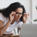 Young woman freelancer with bad eyesight using laptop, using glasses, home interior, copy space. Curly lady holding her glasses and squinting, looking at laptop screen, having vision troubles
