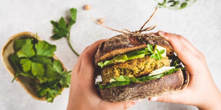 Vegan sandwich with chickpea patty, avocado, cucumber and greens in rye bread in children's hands, top view.