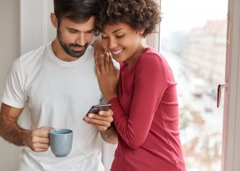 Image of mixed race friendly girl and boy wait for feedback from followers, develop their blog, stand closely near window, drink coffee, chat online, being addicted to smart phones, pose at home