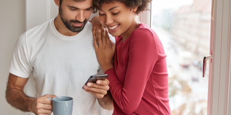 Image of mixed race friendly girl and boy wait for feedback from followers, develop their blog, stand closely near window, drink coffee, chat online, being addicted to smart phones, pose at home