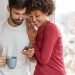 Image of mixed race friendly girl and boy wait for feedback from followers, develop their blog, stand closely near window, drink coffee, chat online, being addicted to smart phones, pose at home