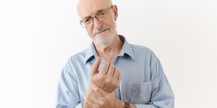 People, age, well being, illness and health problems concept. Studio shot of frustrated upset sixty year old man in spectacles having painful look, rubbing wrist, suffering from pain in joints