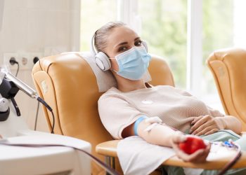 Portrait of young woman in protective mask looking at camera while lying on the couch and receiving a blood transfusion at hospital