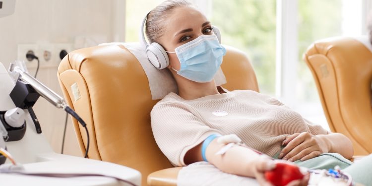 Portrait of young woman in protective mask looking at camera while lying on the couch and receiving a blood transfusion at hospital
