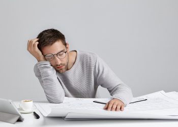 Tired young attractive man sleeps at work place, has much work, being fatigue and exhausted, isolated over white studio background. Sleepy male engineer has deadline, busy all day, has sleppless night