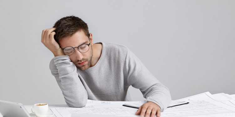 Tired young attractive man sleeps at work place, has much work, being fatigue and exhausted, isolated over white studio background. Sleepy male engineer has deadline, busy all day, has sleppless night