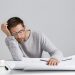 Tired young attractive man sleeps at work place, has much work, being fatigue and exhausted, isolated over white studio background. Sleepy male engineer has deadline, busy all day, has sleppless night
