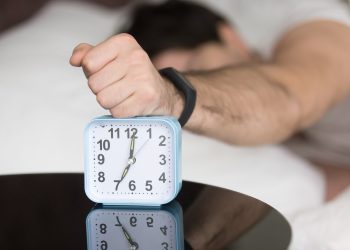 Annoyed sleepy man turning off alarm, hitting button on the clock by his fist after sleepless night, angry and frustrate sleeper feeling not rested and tired, lying in bed and wanting to sleep more