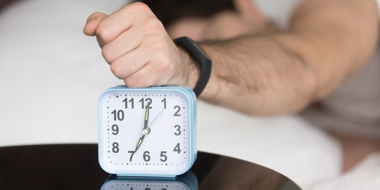 Annoyed sleepy man turning off alarm, hitting button on the clock by his fist after sleepless night, angry and frustrate sleeper feeling not rested and tired, lying in bed and wanting to sleep more
