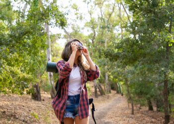 Long-haired woman taking photo of nature and standing on road in forest. Blonde Caucasian lady holding camera and shooting landshate. Backpacking tourism, adventure and summer vacation concept