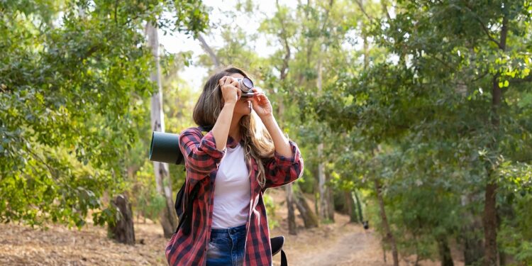 Long-haired woman taking photo of nature and standing on road in forest. Blonde Caucasian lady holding camera and shooting landshate. Backpacking tourism, adventure and summer vacation concept