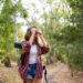 Long-haired woman taking photo of nature and standing on road in forest. Blonde Caucasian lady holding camera and shooting landshate. Backpacking tourism, adventure and summer vacation concept