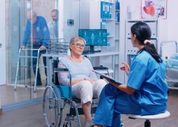 Nurse talking with senior woman with walking disabilities sitting in a wheelchair, into a private modern recovery clinic or hospital. Handicapped old retired patient medical consultation and advice