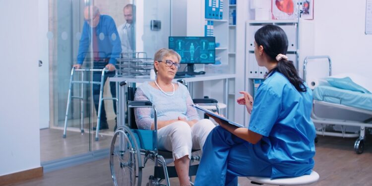 Nurse talking with senior woman with walking disabilities sitting in a wheelchair, into a private modern recovery clinic or hospital. Handicapped old retired patient medical consultation and advice