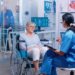 Nurse talking with senior woman with walking disabilities sitting in a wheelchair, into a private modern recovery clinic or hospital. Handicapped old retired patient medical consultation and advice