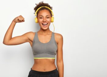 Horizontal view of dark skinned woman in good mood, raises arm with muscles, has strong body, dressed in gym outfit, listens audio via modern headphones, poses indoor. Fitness and music concept