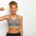 Horizontal view of dark skinned woman in good mood, raises arm with muscles, has strong body, dressed in gym outfit, listens audio via modern headphones, poses indoor. Fitness and music concept