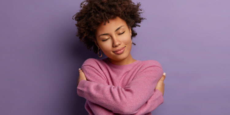 Romantic woman embraces herself, keeps arms crossed over body, touches shoulders, enjoys coziness, wears sweater, tilts head, has high self esteem, loves herself, isolated on purple background