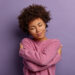 Romantic woman embraces herself, keeps arms crossed over body, touches shoulders, enjoys coziness, wears sweater, tilts head, has high self esteem, loves herself, isolated on purple background