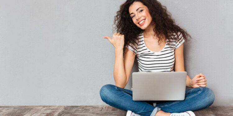 Nice woman with beautiful smile sitting in lotus pose on the floor with silver computer on legs gesturing thumb aside, submitting something copy space