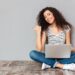 Nice woman with beautiful smile sitting in lotus pose on the floor with silver computer on legs gesturing thumb aside, submitting something copy space